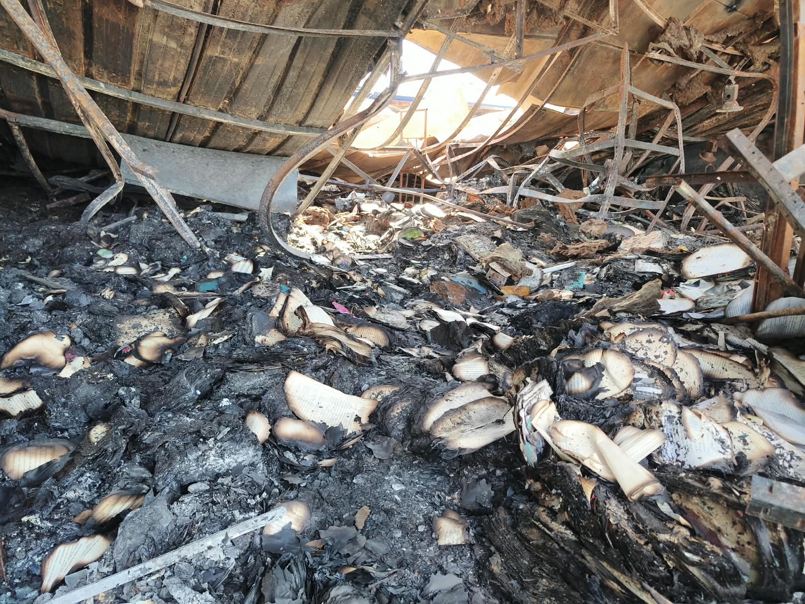 Inside a burned-out book stall: the corrugated metal roof has collapsed onto buckled shelving frames, daylight showing through a hole in it. The floor is a thick layer of ash and charred books, their pale page edges still fanning out of the blackened stacks.